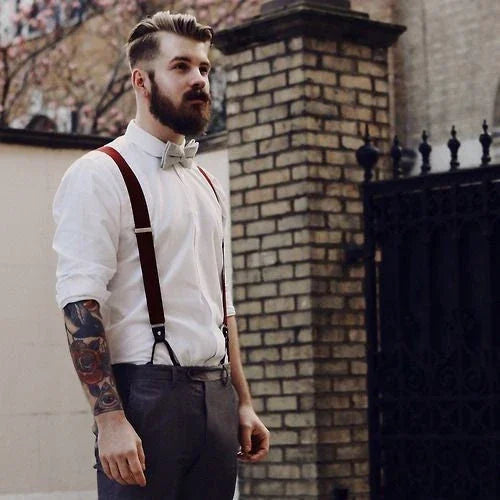 Hombre con pajarita gris y tirantes rojos, camisa blanca y pantalón gris, estilo moderno con tatuajes