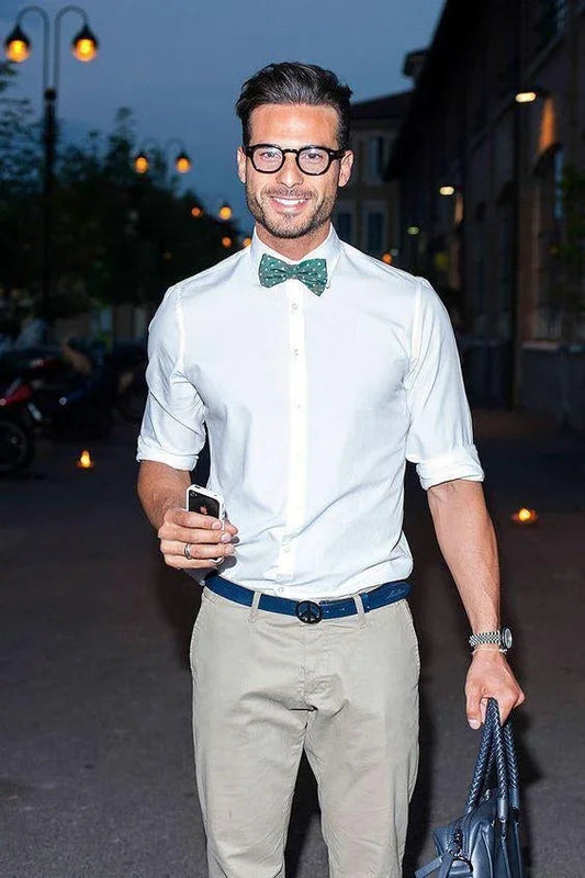 Hombre joven con gafas y pajarita verde con puntos blancos, camisa blanca y pantalones beige, en exterior nocturno