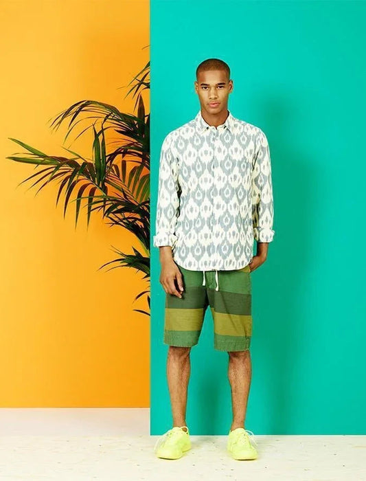 Hombre joven con camisa blanca estampada, bermudas verdes y zapatillas amarillas, fondo bicolor y planta tropical