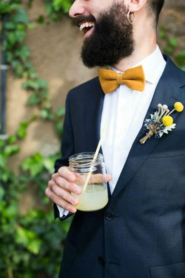 Hombre con pajarita mostaza y traje azul oscuro, con flor en solapa y vaso con bebida y pajilla