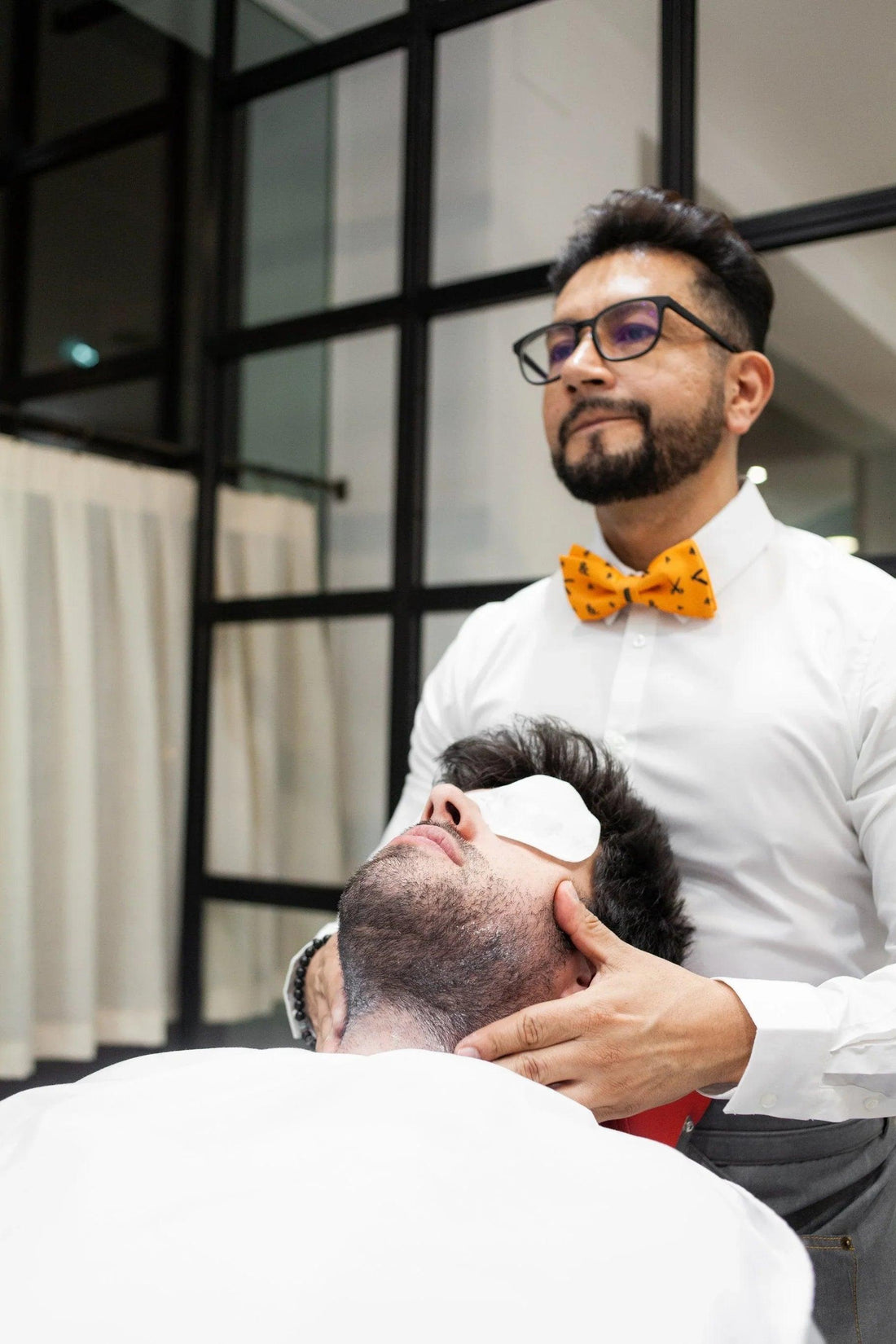 Barbero con pajarita naranja atendiendo a cliente con toalla en los ojos en barbería moderna