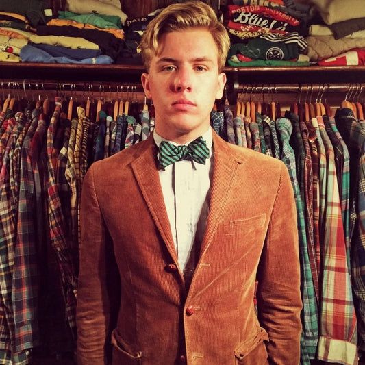 Hombre joven con pajarita verde de rayas, camisa blanca y americana marrón frente a armario con camisas a cuadros