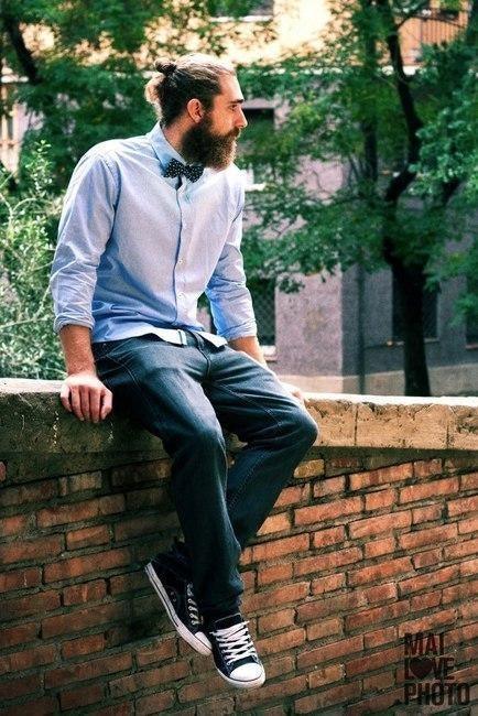 Hombre con pajarita original y camisa azul claro sentado sobre muro de ladrillos en ambiente urbano con árboles