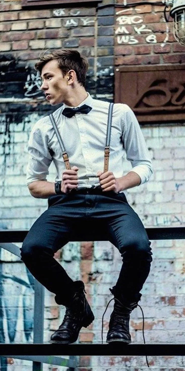 Hombre joven con pajarita negra y tirantes, estilo rockabilly, sentado frente a pared de ladrillo con grafitis