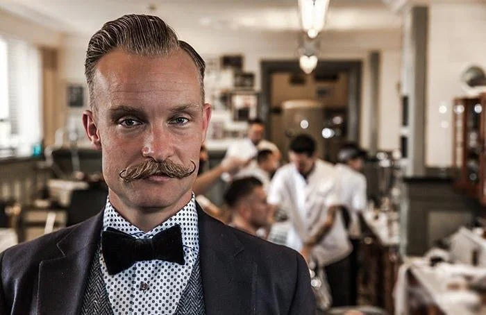 Hombre vestido con pajarita negra y camisa de lunares en barbería clásica con estilistas al fondo
