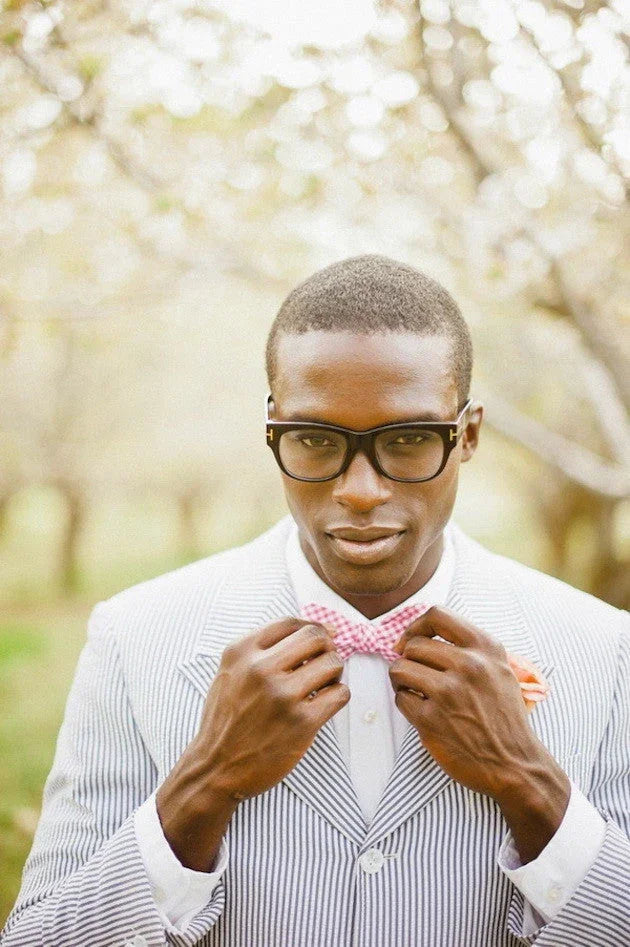 Hombre con gafas ajustando pajarita rosa a cuadros en traje con rayas finas en fondo de árboles floridos