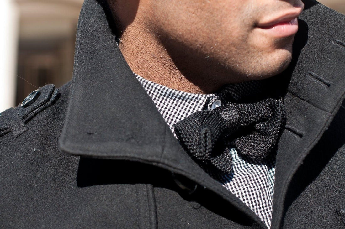 Hombre con abrigo negro usando camisa a cuadros y pajarita negra de punto artesanal en detalle